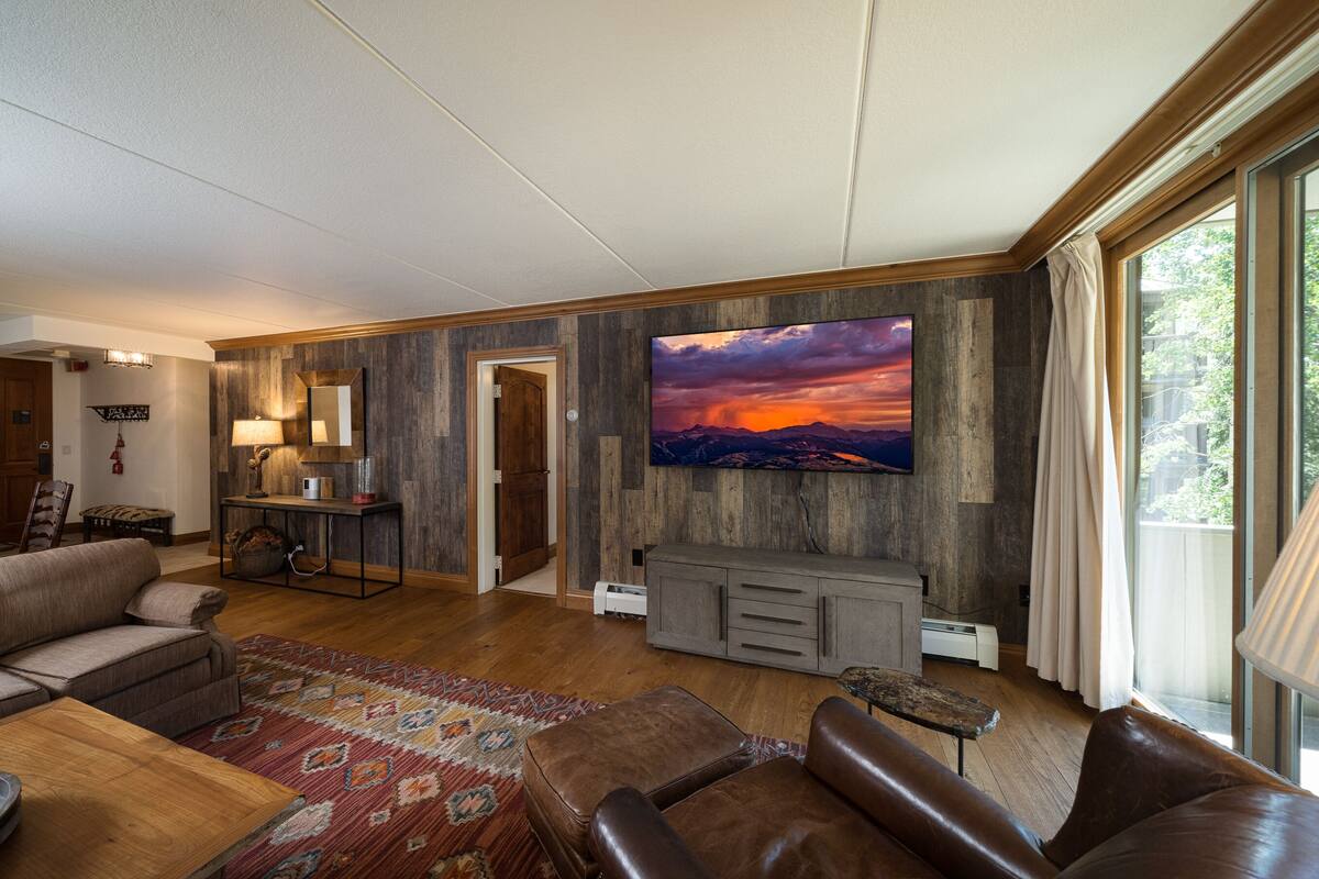 A spacious living room with a large flat-screen TV, leather sofas, a wooden coffee table, and a patterned rug.