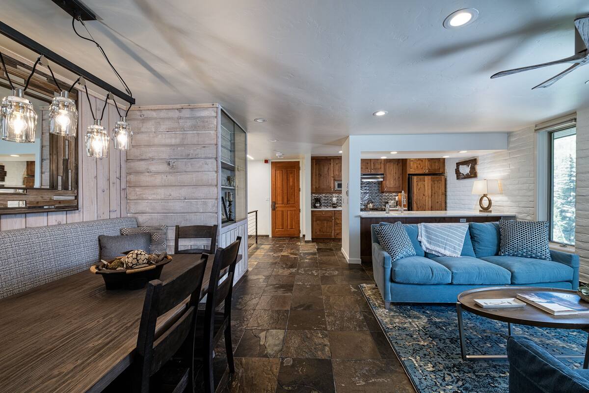 A modern kitchen with wooden cabinets, a dining area with a table and chairs, and a living room with a blue sofa and a coffee table.
