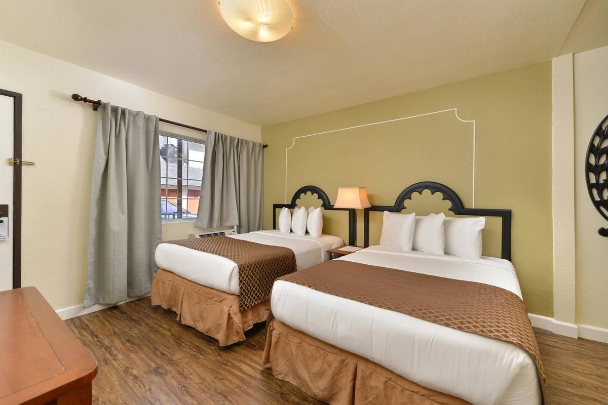 A hotel room with two beds, a wooden floor, a window with curtains, and a wall decoration.
