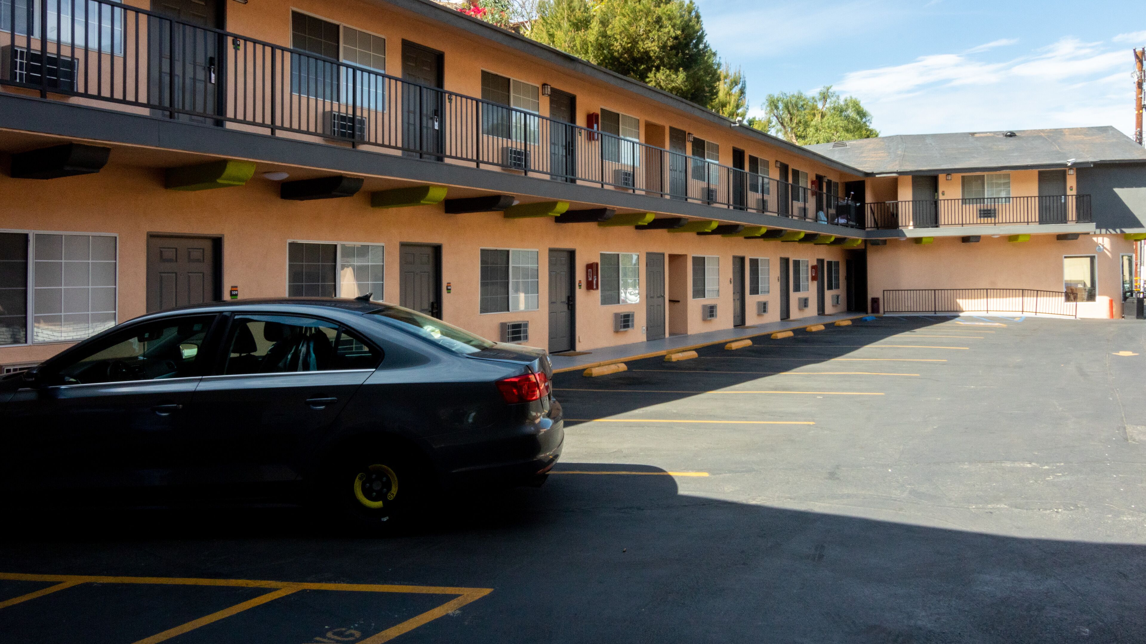 A parking lot with a black car parked in front of a motel with multiple balconies and a flat-roofed building.