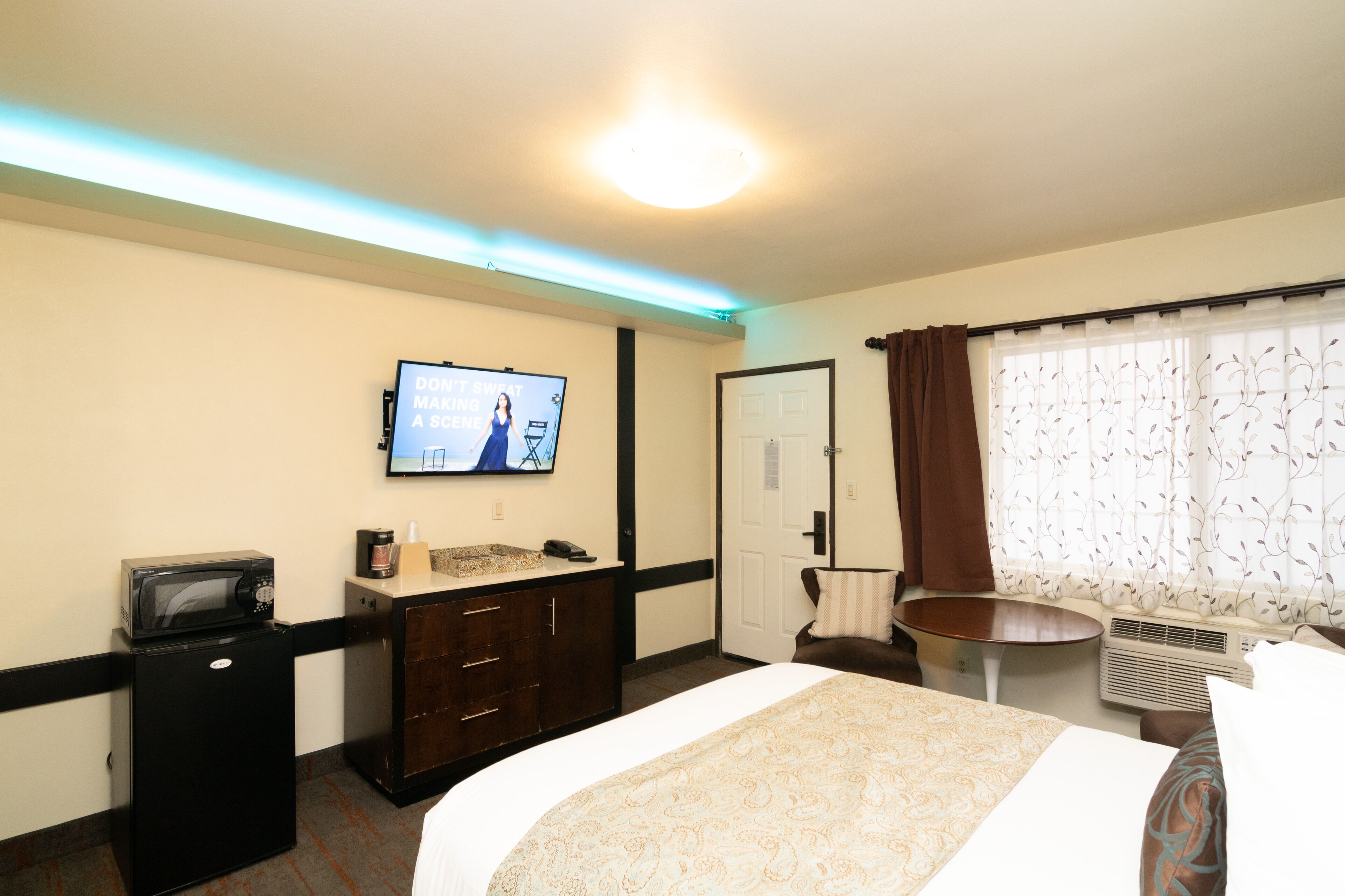 Une chambre d’hôtel équipée d’un lit, d’un téléviseur fixé au mur, d’un petit réfrigérateur, d’un bureau et d’une fenêtre avec des rideaux.