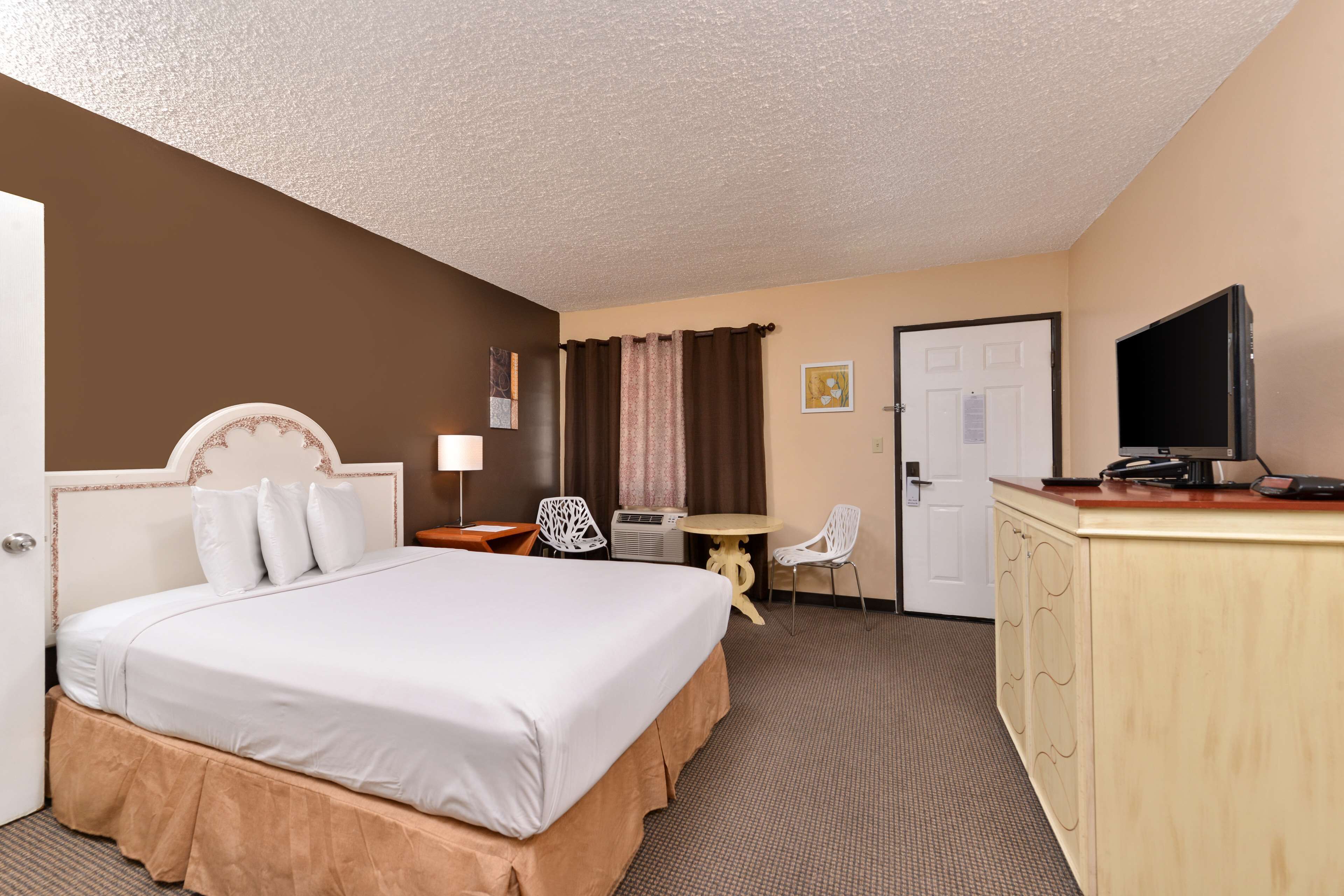 A hotel room with a large bed, a desk with a television, a chair, a small table, and a door.