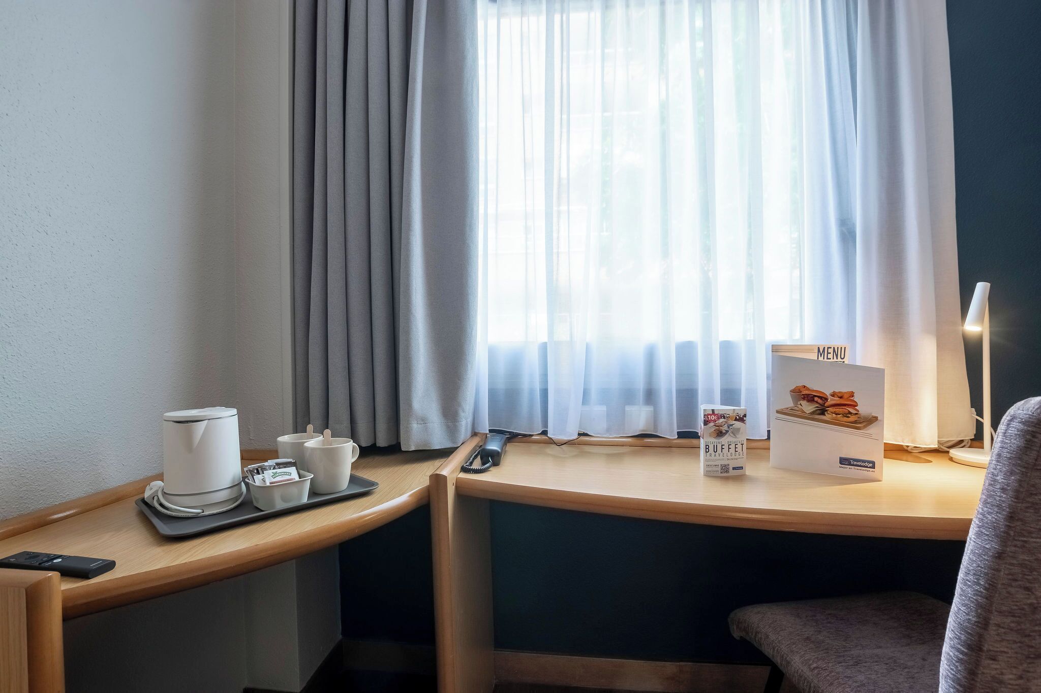 Habitación de hotel con escritorio, una bandeja con cafetera, tazas y un control remoto. Hay un cartel con el menú en el escritorio y una lámpara en el lado derecho.