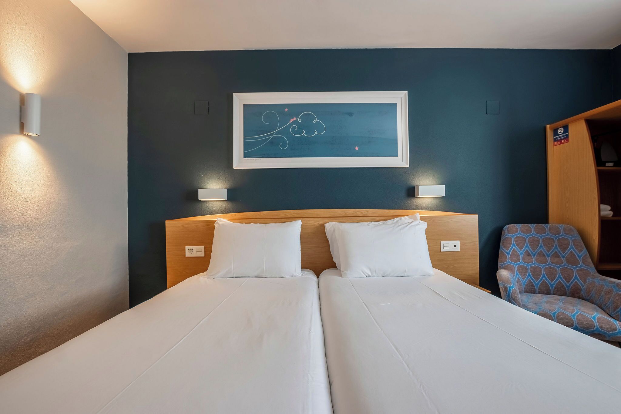 Habitación de hotel con dos camas individuales, cabeceros, lámparas de noche, una obra de arte enmarcada y un sillón estampado.