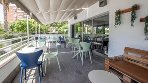 Een terras met tafels en stoelen, een houten bankje en uitzicht op gebouwen en bomen.