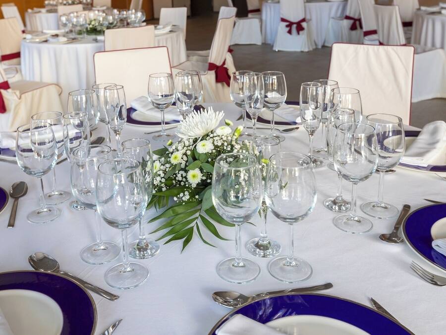 Una mesa de banquete dispuesta con varias copas de vino, platos y cubiertos, rodeada de sillas con lazos blancos y rojos.