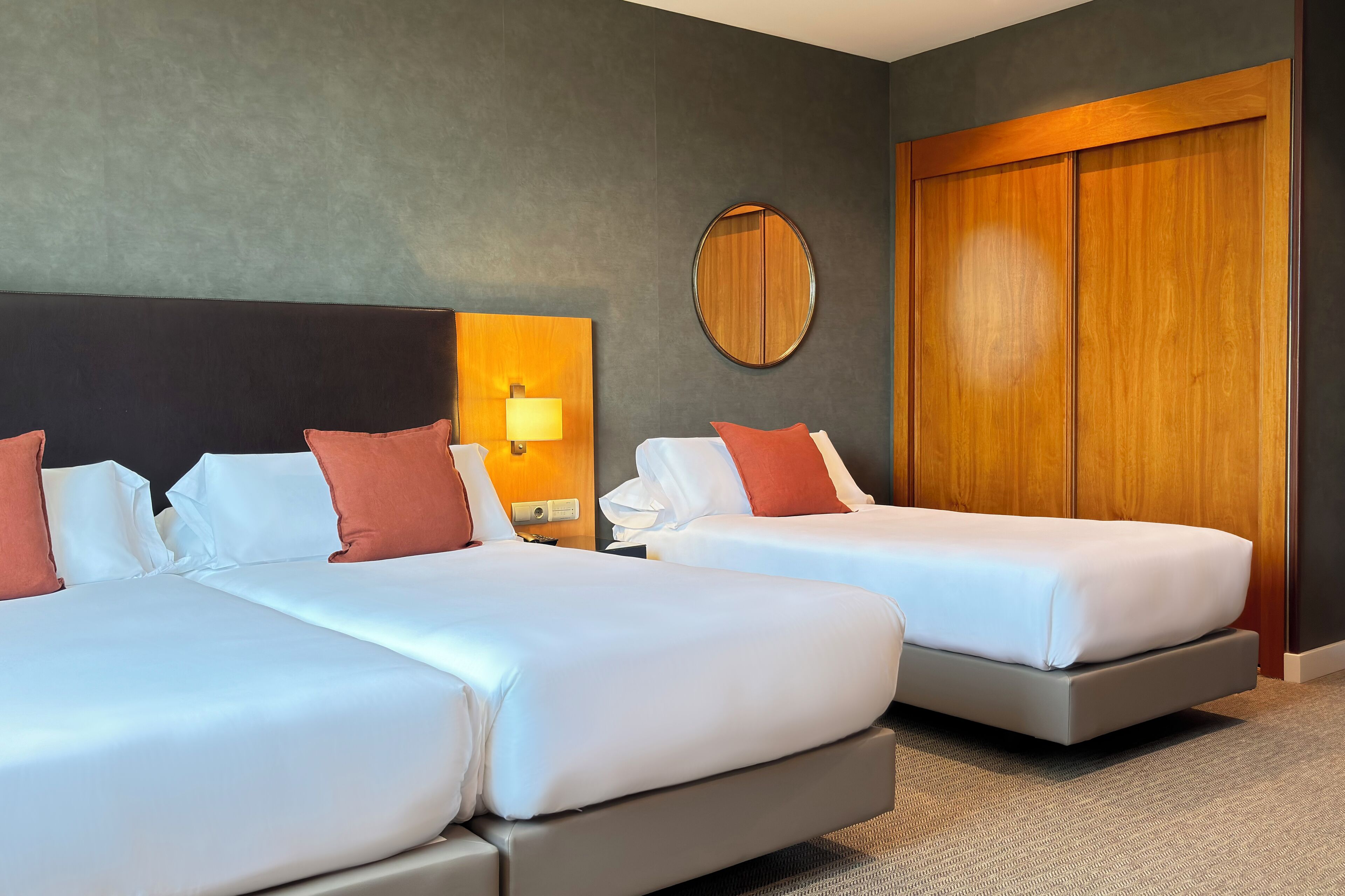 A hotel room with two beds, a round mirror, a wooden wardrobe, and a nightstand with a lamp.