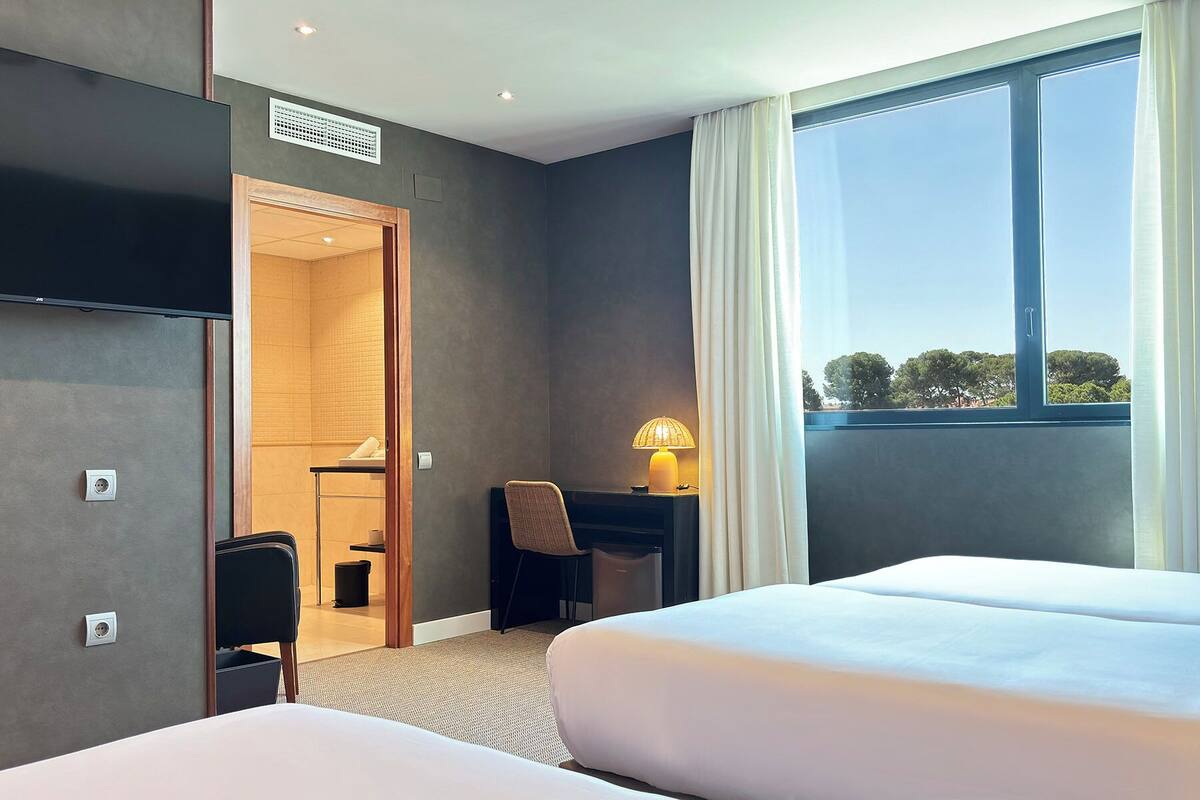 A hotel room with two beds, a desk, a chair, a lamp, and a large window with a view of trees.