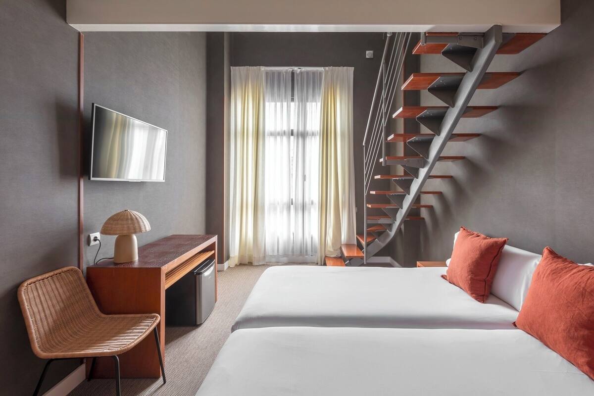 A modern hotel room with a bed, a desk, a chair, a TV, and a staircase.
