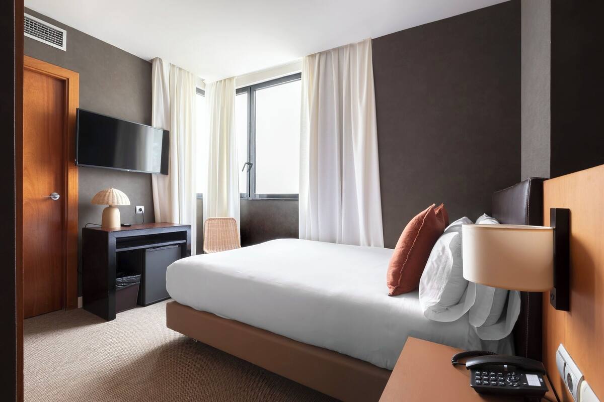 A hotel room with a bed, a desk, a TV, a lamp, a phone, and a window with curtains.