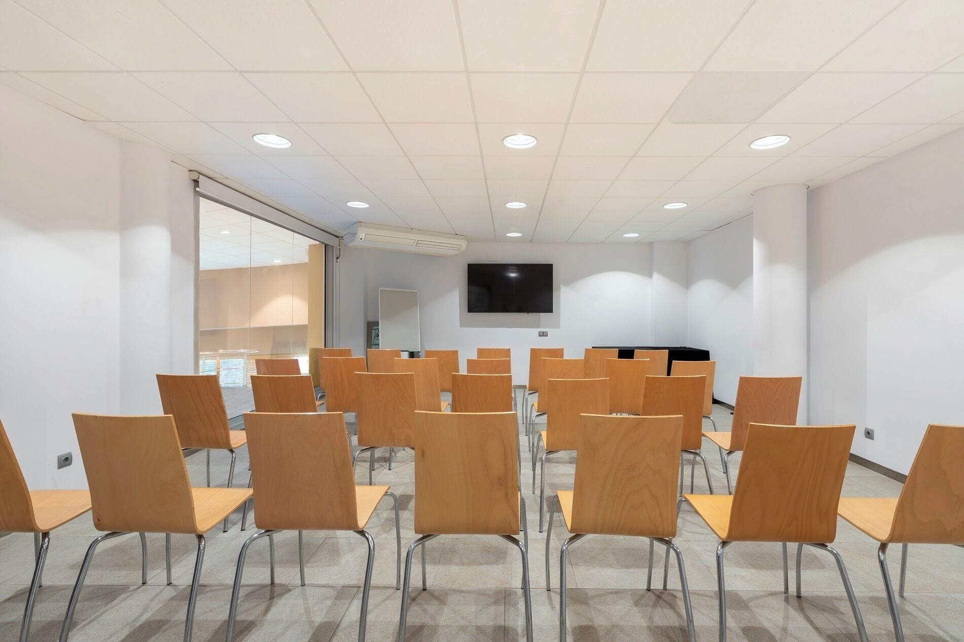 Un salón de conferencias con filas de sillas de madera, un televisor de pantalla plana montado en la pared y un panel de vidrio en un lateral.