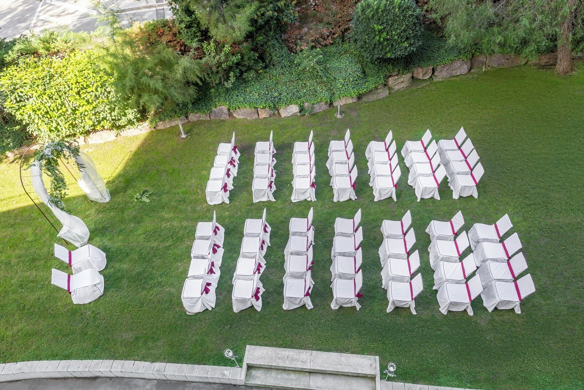 Un espace extérieur aménagé avec des rangées de chaises et de tables blanches, entouré d’un mur de pierre et de verdure.
