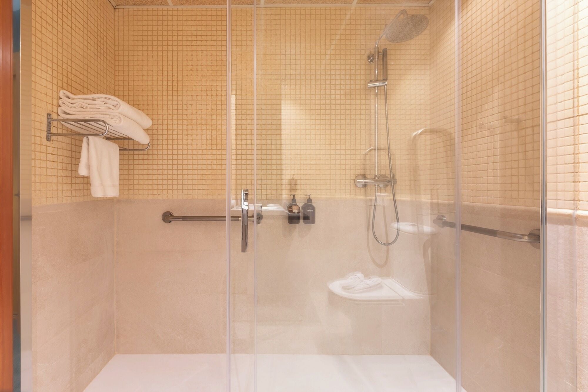 Een douchecabine met glazen wanden, een betegelde muur, een handdoekhouder met opgevouwen handdoeken en een handdouche.