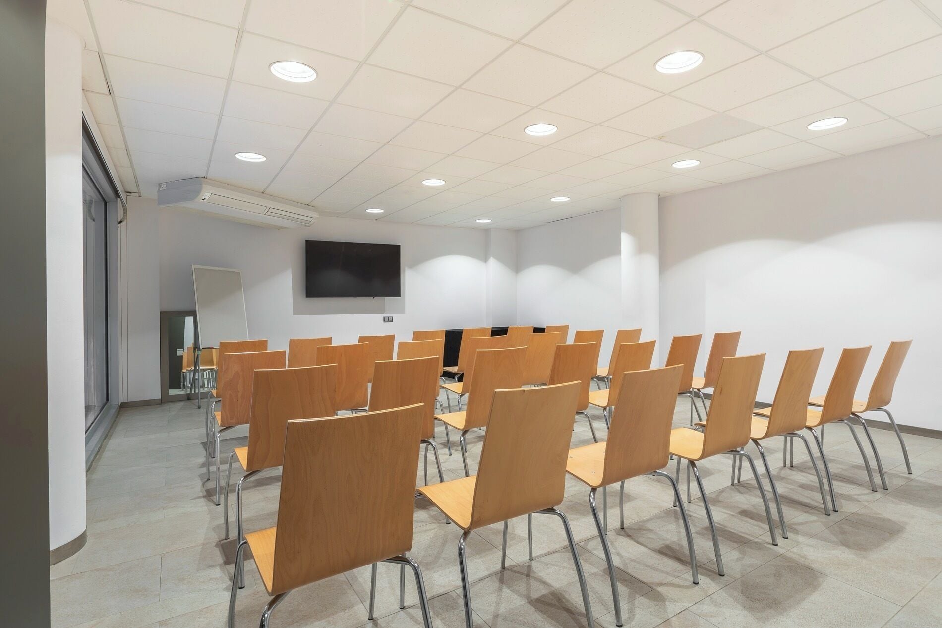 Un salón de conferencias con filas de sillas de madera, un televisor de pantalla plana en la pared y suelo de baldosas.