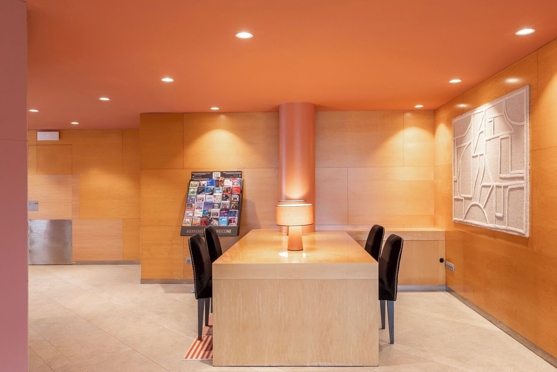 Un moderno vestíbulo de hotel con un mostrador de recepción de madera en el centro, un gran panel informativo en la pared y iluminación empotrada en el techo.