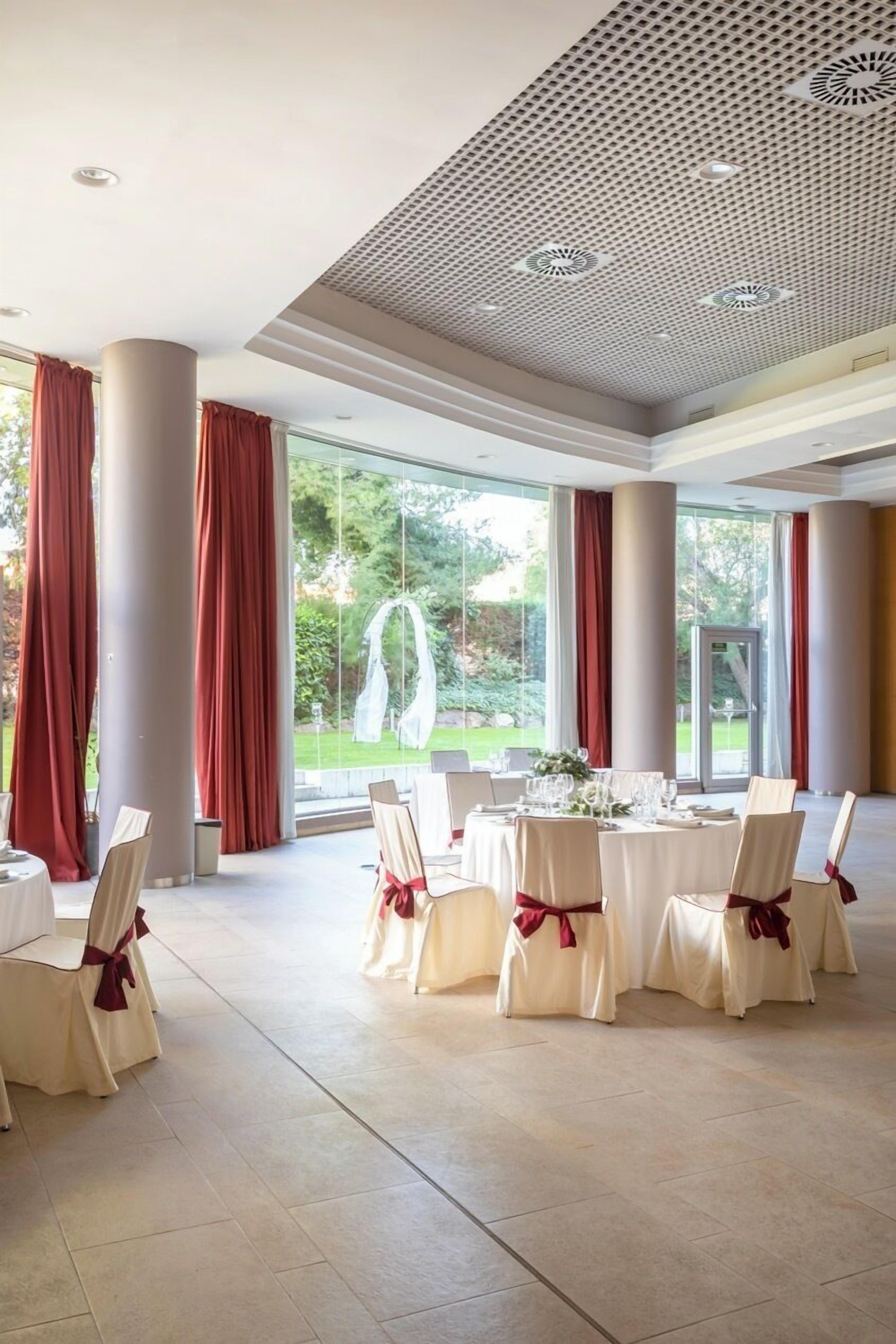 A spacious indoor venue with large windows, red curtains, and a high ceiling with a geometric pattern. Tables are set with white tablecloths and chairs with red sashes.