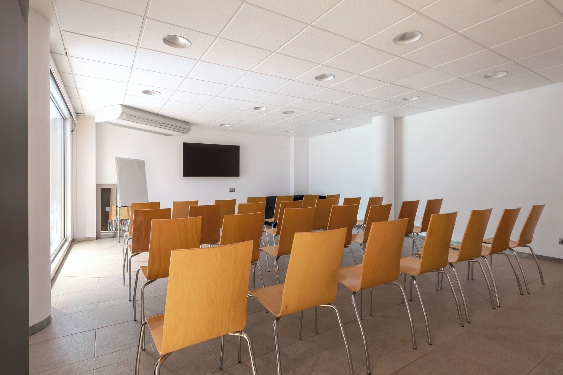 Un salón de conferencias con filas de sillas de madera, un televisor de pantalla plana en la pared y un amplio ventanal que proporciona luz natural.