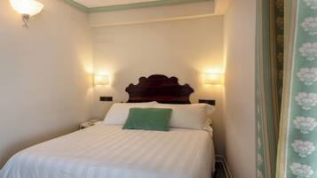 Cama bem arrumada com lençóis brancos e um travesseiro verde, com duas luminárias fixadas na parede.
