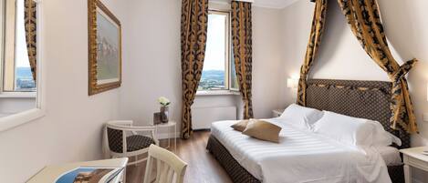 A hotel room with a large bed, a desk with a chair, a small table with a vase, and a window with curtains.