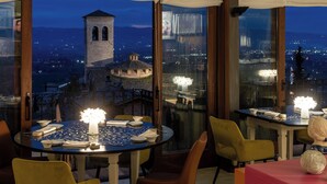 A dining area with tables and chairs, a view of a cityscape, and a tower in the background. - Hotel Fontebella (Assisi)