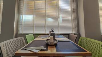 Un comedor con una mesa dispuesta para cuatro personas, rodeada de sillas verdes, y una ventana con persianas al fondo.