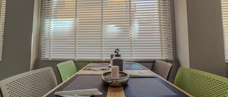 Un comedor con una mesa dispuesta para cuatro personas, rodeada de sillas verdes, y una ventana con persianas al fondo.