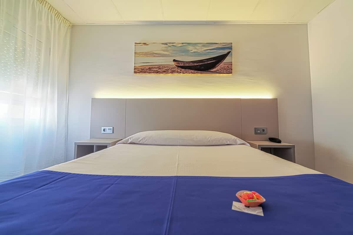 Una habitación de hotel con cama, mesita de noche, lámpara y un cuadro de un barco en la playa.