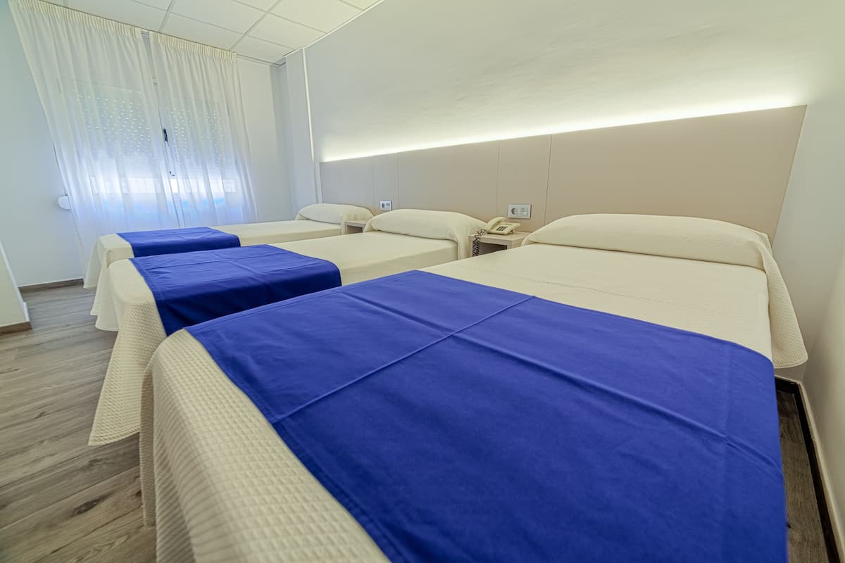 A hotel room with three beds, each with a blue bedspread and white pillows. The room has a wooden floor and white walls.