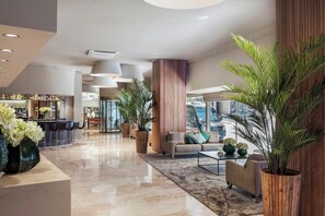 Un hall d’hôtel moderne avec un bar, des espaces pour s’asseoir et de grandes plantes en pot.