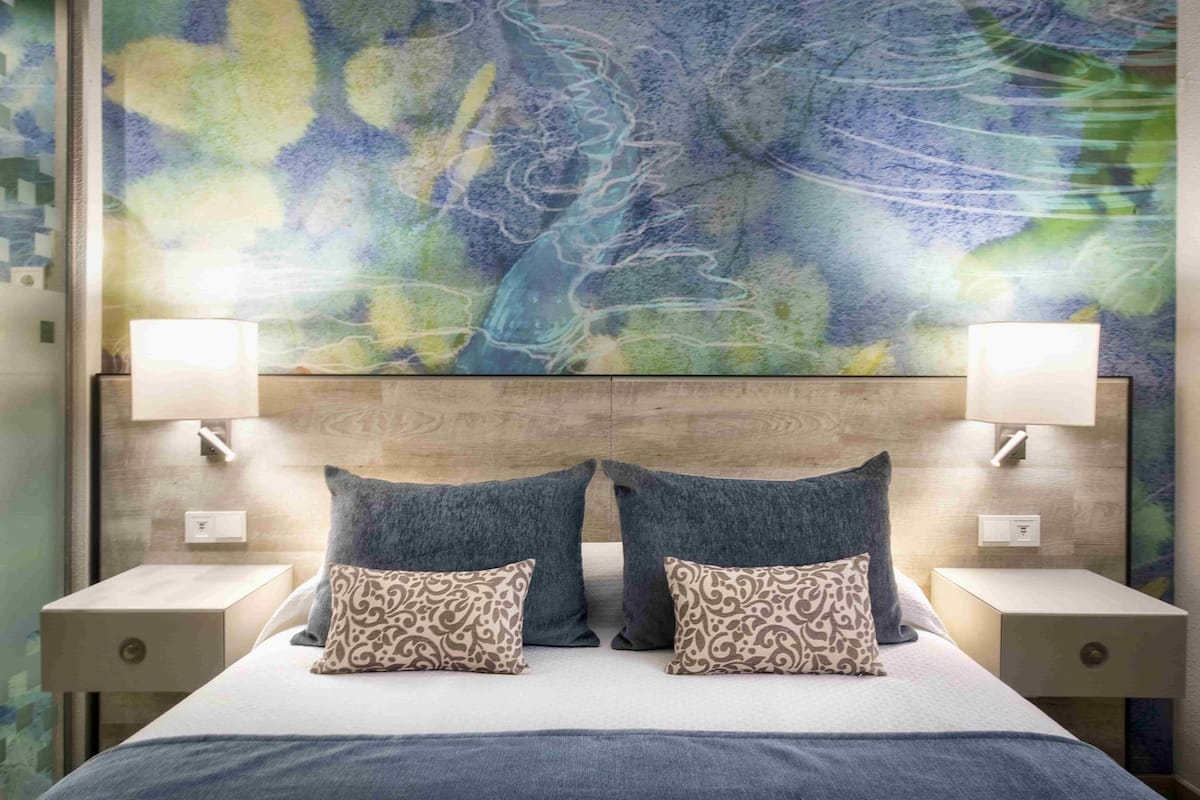 Una habitación de hotel moderna con un cuadro abstracto grande en la pared, un cabecero de madera, dos mesitas de noche con lámparas y una cama con almohadones estampados.