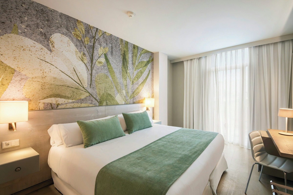 A hotel room with a large bed, a desk, and a chair. There is a painting on the wall and a window with curtains.