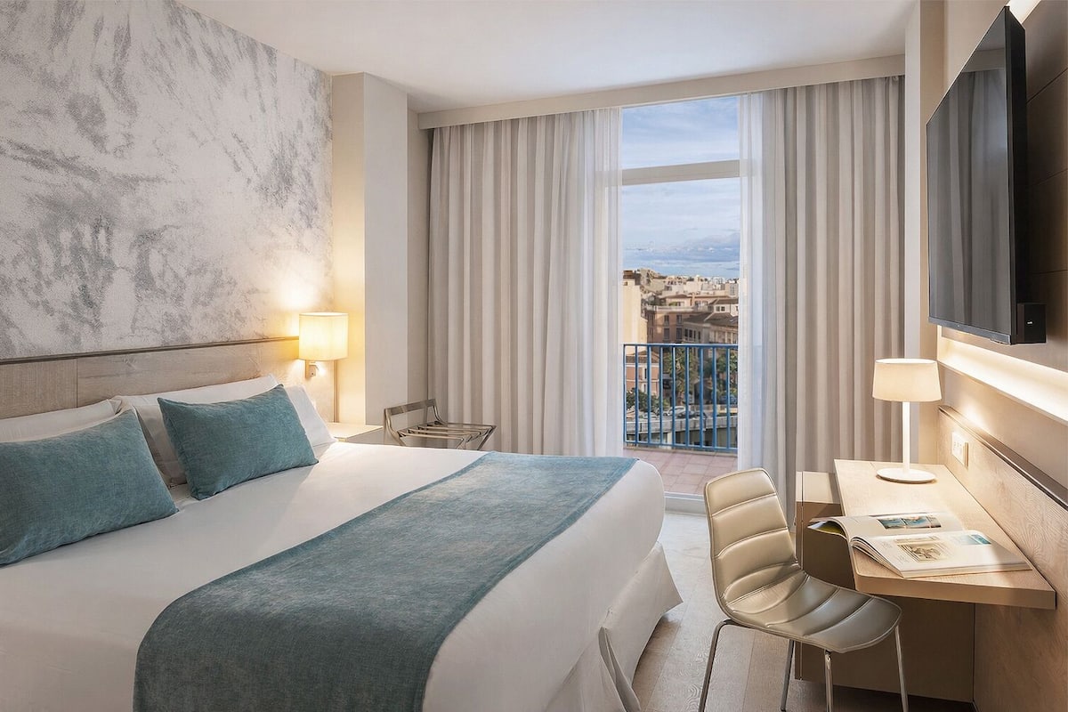 Habitación de hotel con una cama grande, un escritorio con silla, televisión y balcón con cortinas.