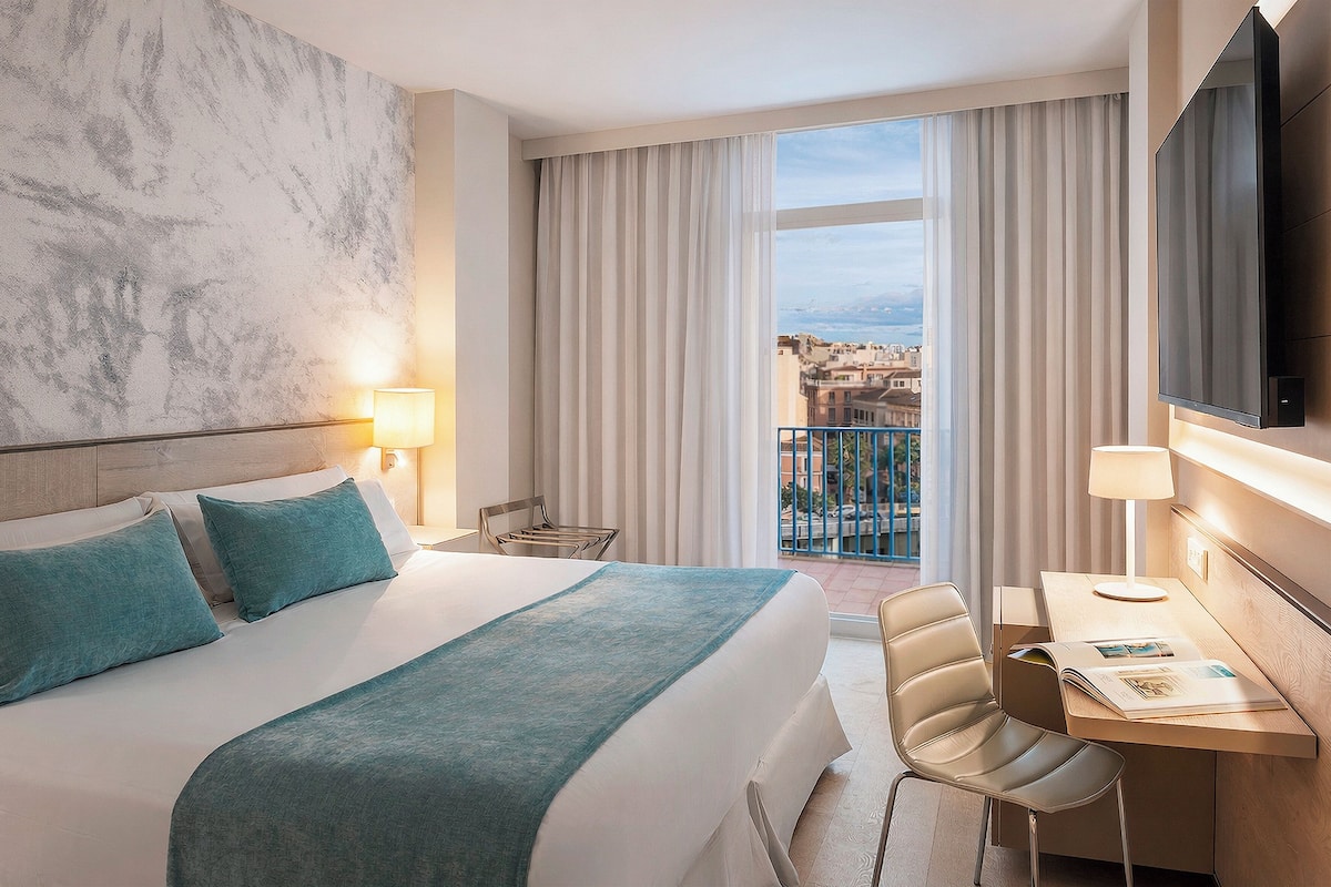 Una habitación de hotel con cama, escritorio, silla, televisor y vista a la ciudad.