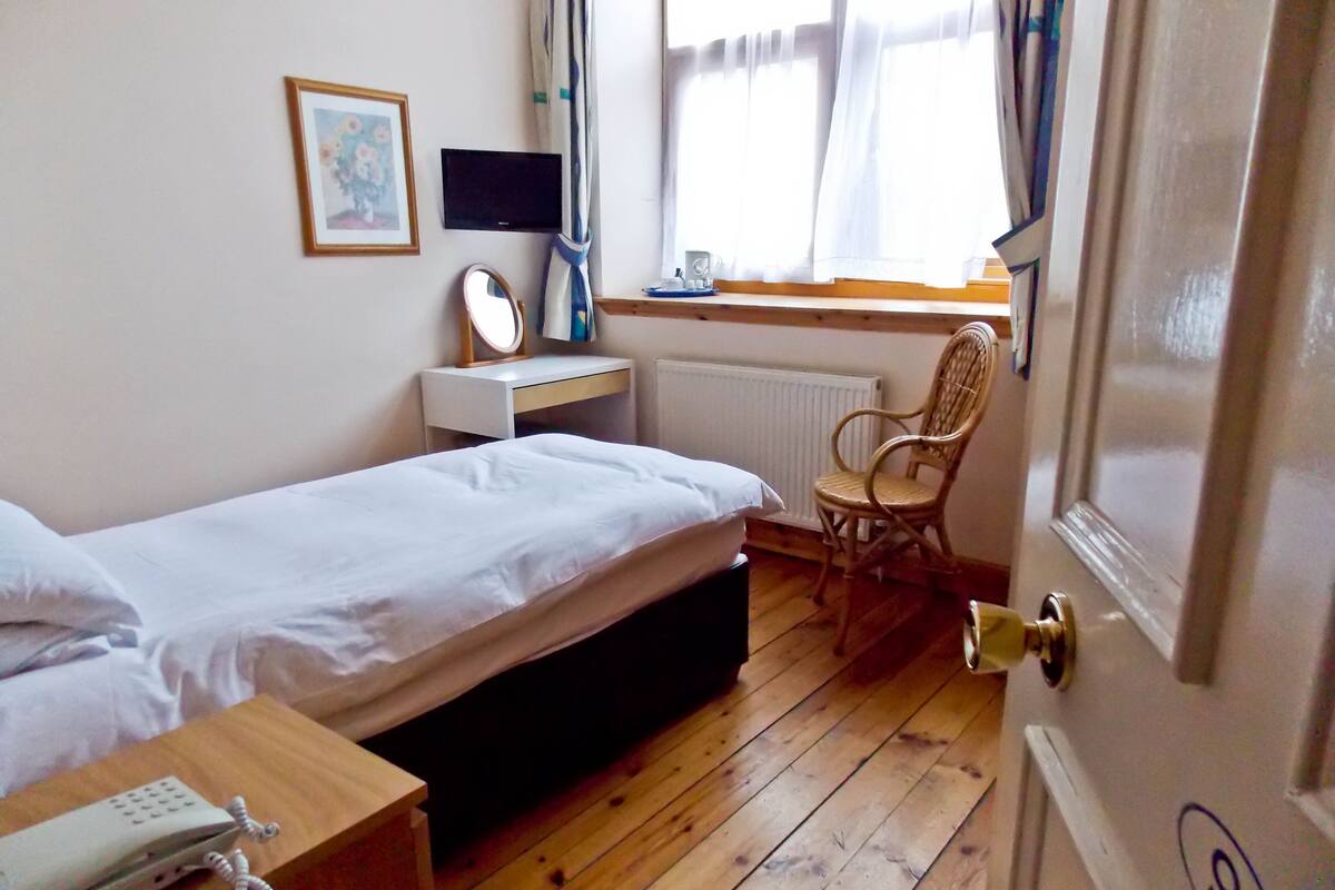 A bedroom with a single bed, a bedside table with a phone, a small desk with a mirror, a chair, and a window with curtains.