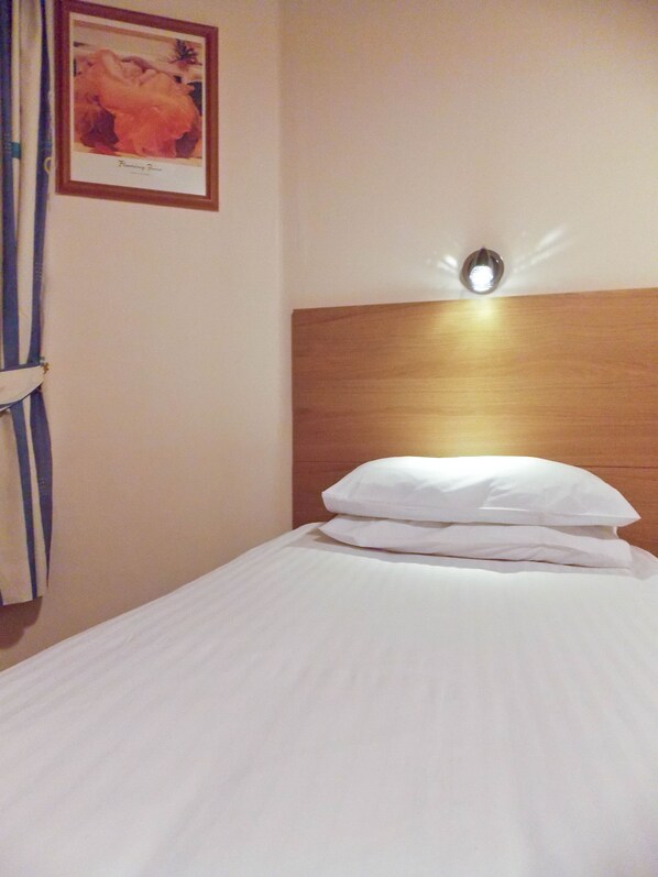 A hotel room with a neatly made bed, a wooden headboard, a framed picture on the wall, and a light fixture above the bed.