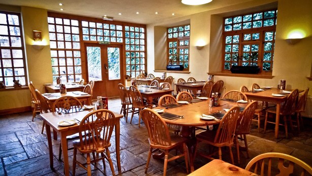 A spacious dining area with multiple wooden tables and chairs, large windows with a view of trees, and a fire exit sign. - The Victorian House (Glasgow)
