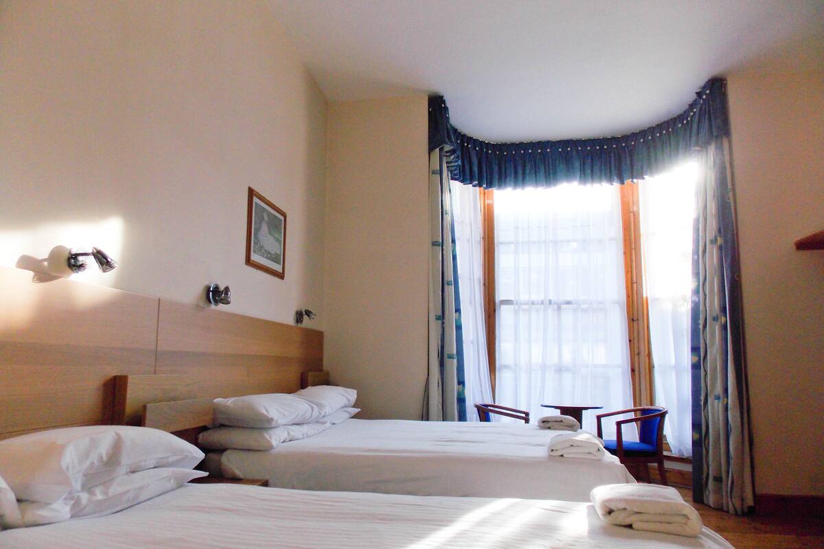 A hotel room with two beds, a wooden headboard, a small table, a chair, and a window with curtains.