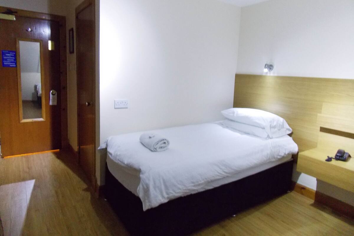 A small, neatly made bed with a white duvet and pillows, a wooden nightstand with a lamp, and a wooden floor.