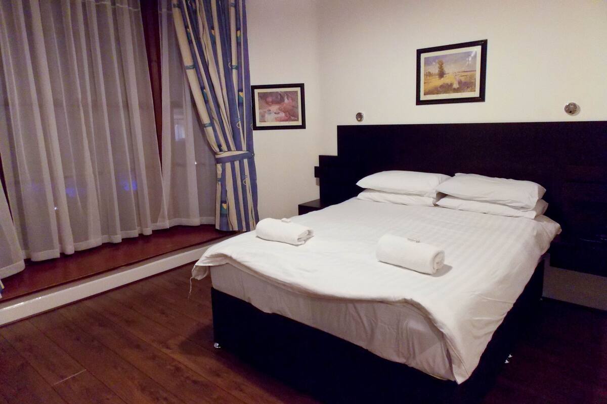 A neatly made bed with white linens and two rolled towels, a dark headboard, and a window with sheer curtains and a striped drape.