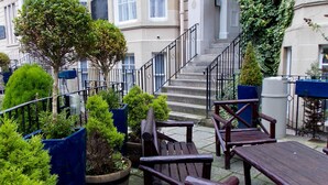 A courtyard with a wooden bench, potted plants, and a staircase leading to a building with large windows and a stone facade. - The Victorian House (Glasgow)