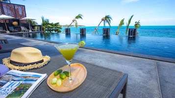 Une scène de bord de piscine : une boisson, un chapeau et un magazine sont posés sur une table, avec vue sur une piscine bordée de palmiers et un ciel dégagé.