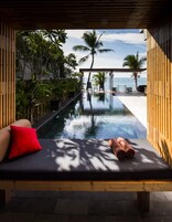 Un salon au bord de la piscine avec une terrasse en bois, une chaise longue agrémentée d’un coussin rouge et une pile de serviettes enroulées.