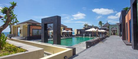 Zona junto a la piscina con sillones de descanso y sombrillas, cielo azul despejado y vista al mar.
