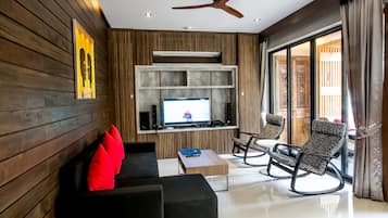 A modern living room with a flat-screen TV, wooden panel walls, a ceiling fan, and a balcony with patterned chairs.