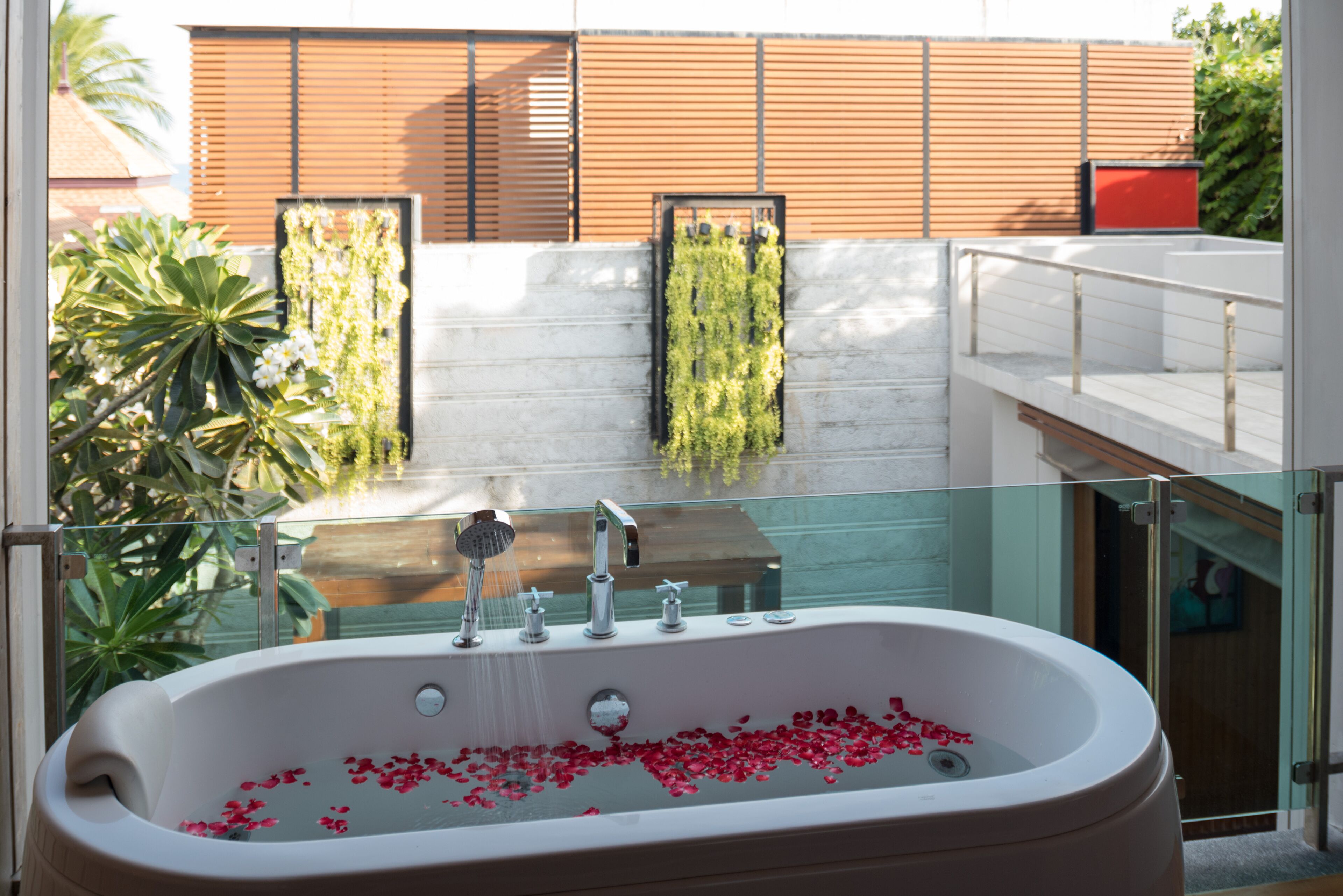A modern bathroom with a large bathtub filled with rose petals, a glass-enclosed shower area, and a view of a building with wooden paneling and greenery.