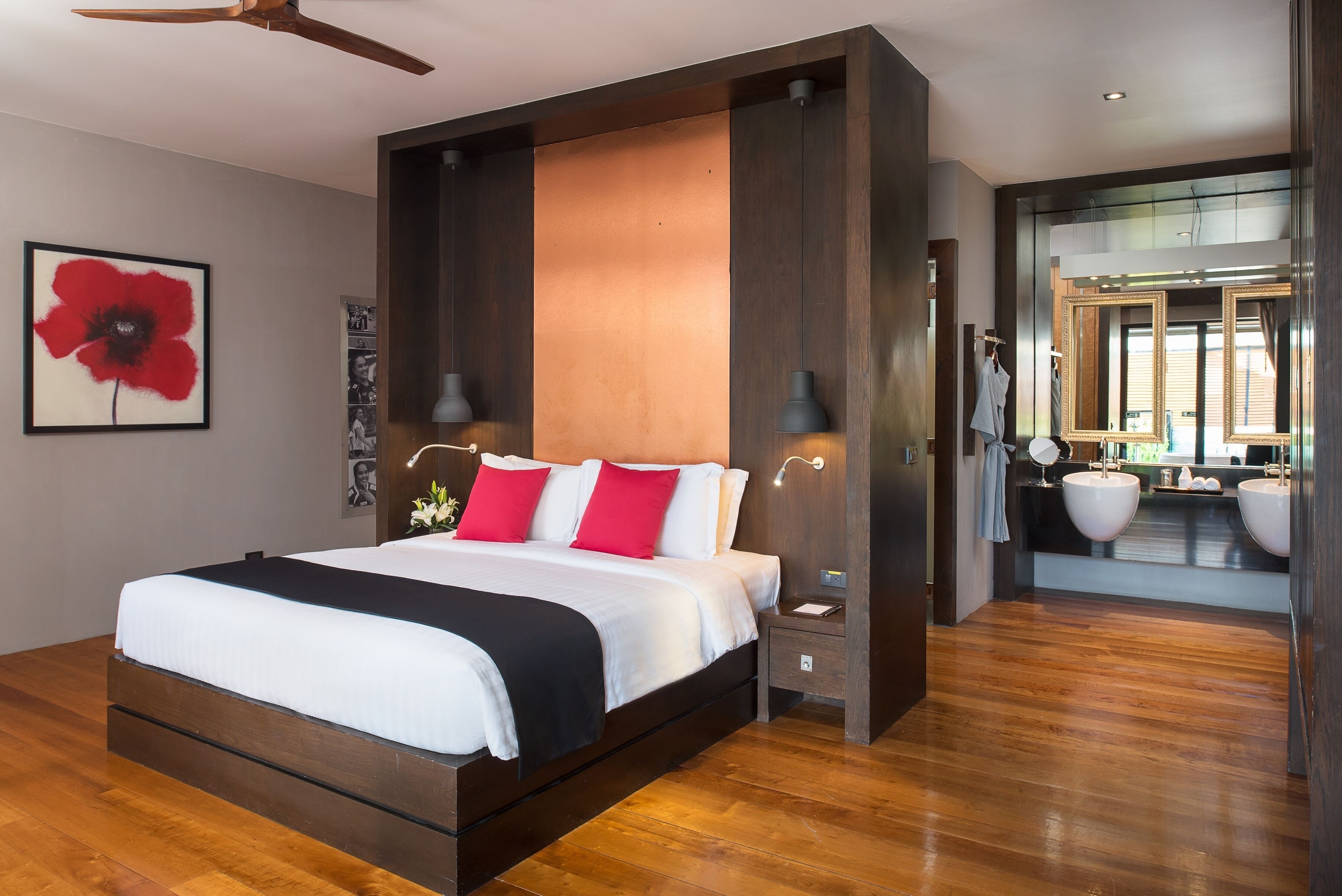 A modern hotel room with a large bed, wooden flooring, a ceiling fan, and a bathroom with two sinks and a mirror.