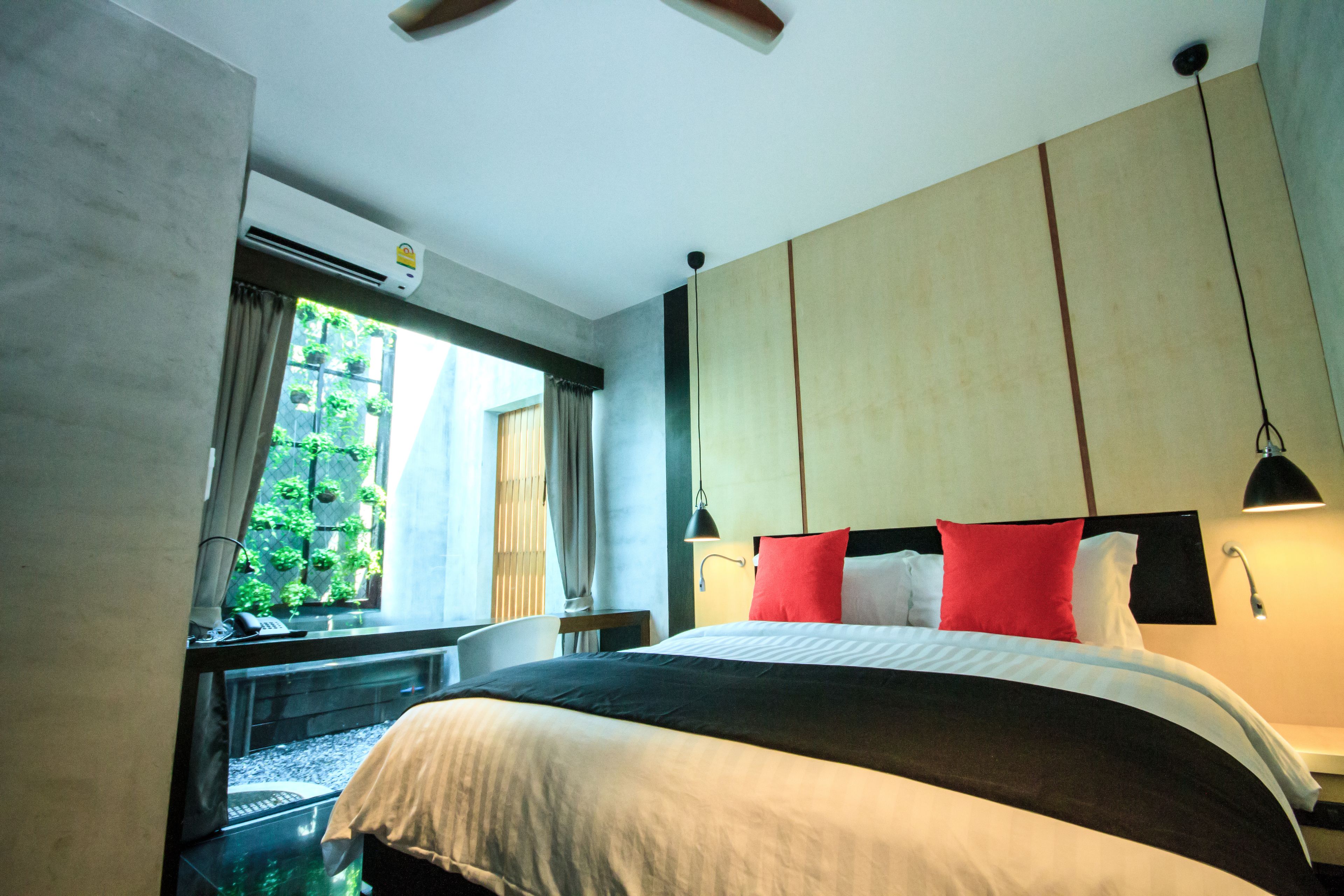 A hotel room with a bed, a window with curtains, an air conditioning unit, and a view of greenery outside.
