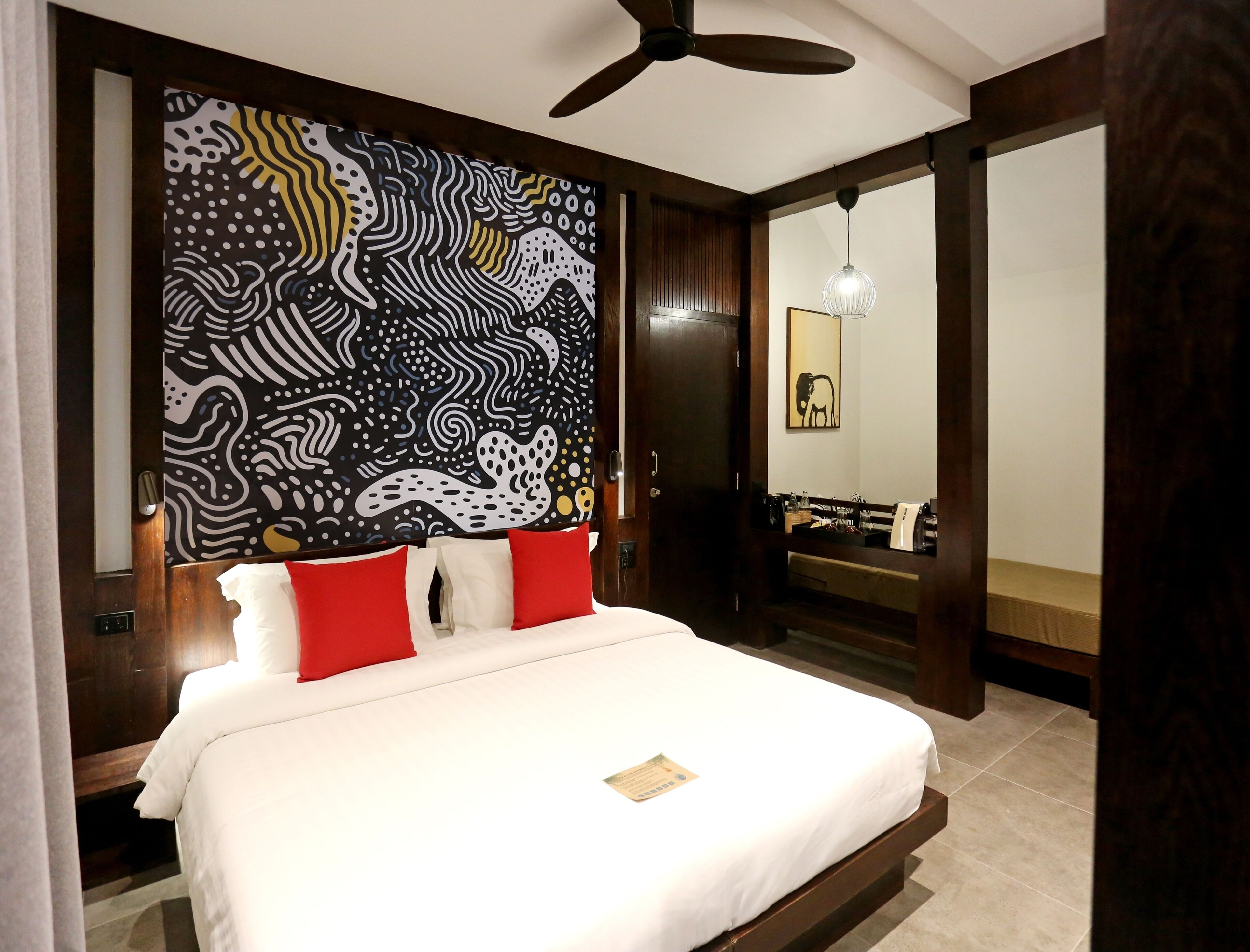 Een hotelkamer met een bed, een plafondventilator, een muur met een zwart-wit abstract patroon en een badkamer zichtbaar door een geopende deur.