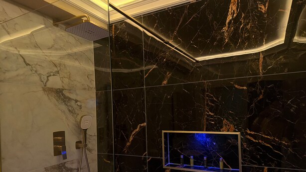 Un baño con ducha, tocador con puerta de vidrio y pared con azulejos de mármol.
