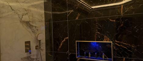Un baño con ducha, tocador con puerta de vidrio y pared con azulejos de mármol.
